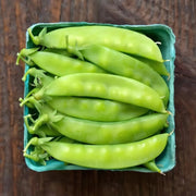 A green cardboard box filled with fresh, green snap peas sits on a wooden surface.