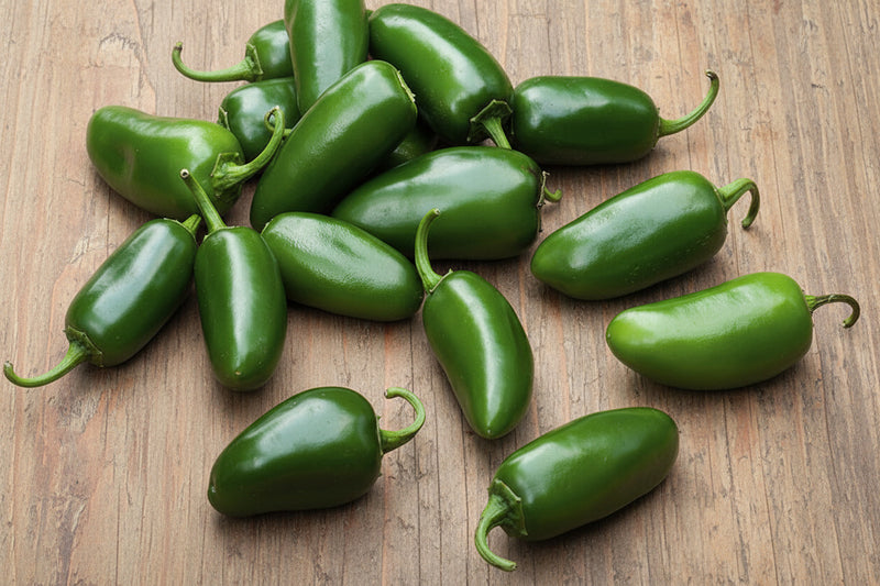 a collection of green jalapeno peppers on a wooden surface, with some of the peppers spilling out of a basket.